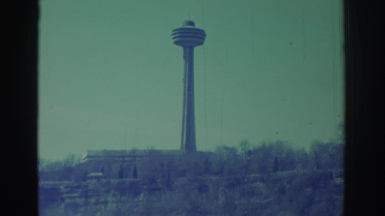 1971 8mm Niagara Falls travel view with an observation tower above bare trees and cliffside, framed by a window.