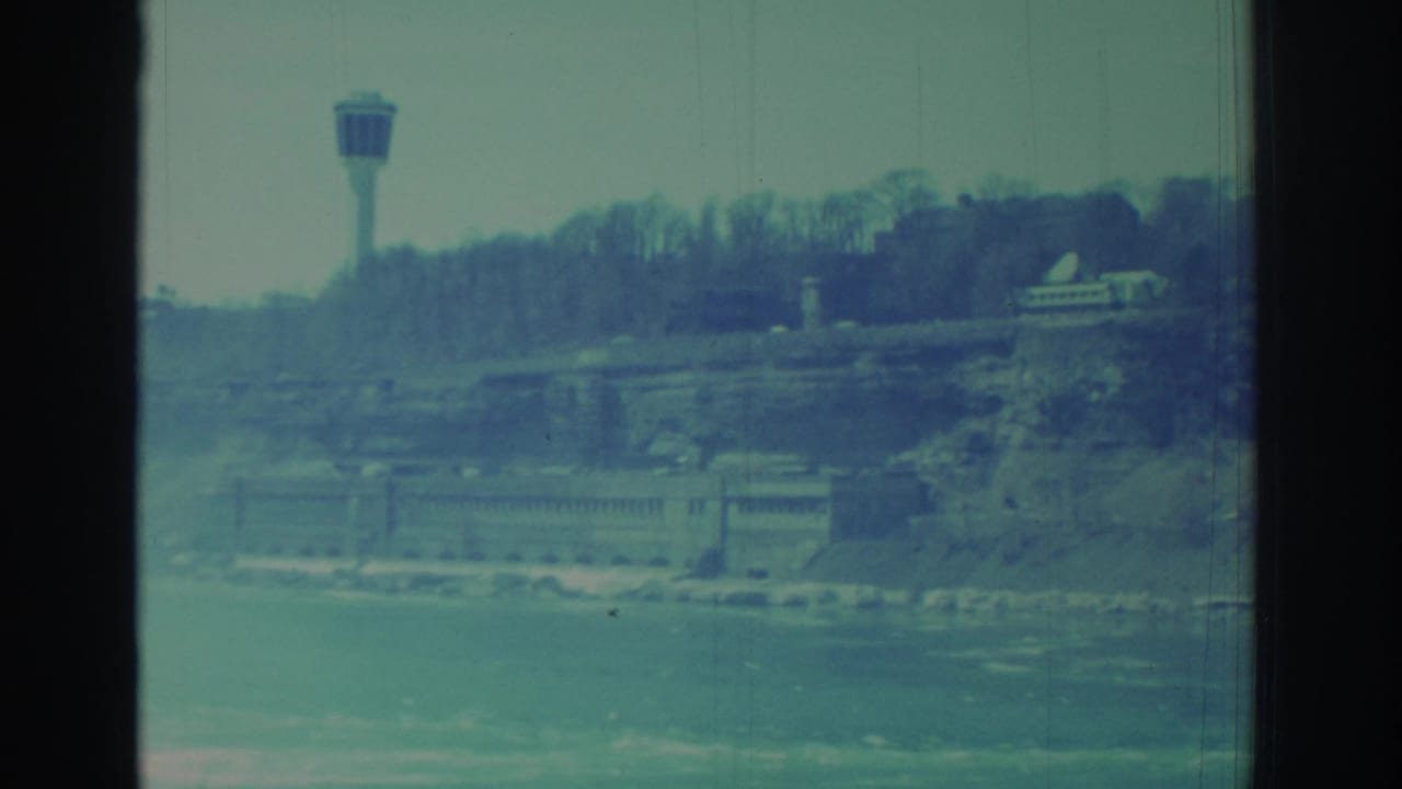 1971 Niagara Falls view across water toward a tower, wooded ridge, and large riverside structure, with 8mm grain and scratches.