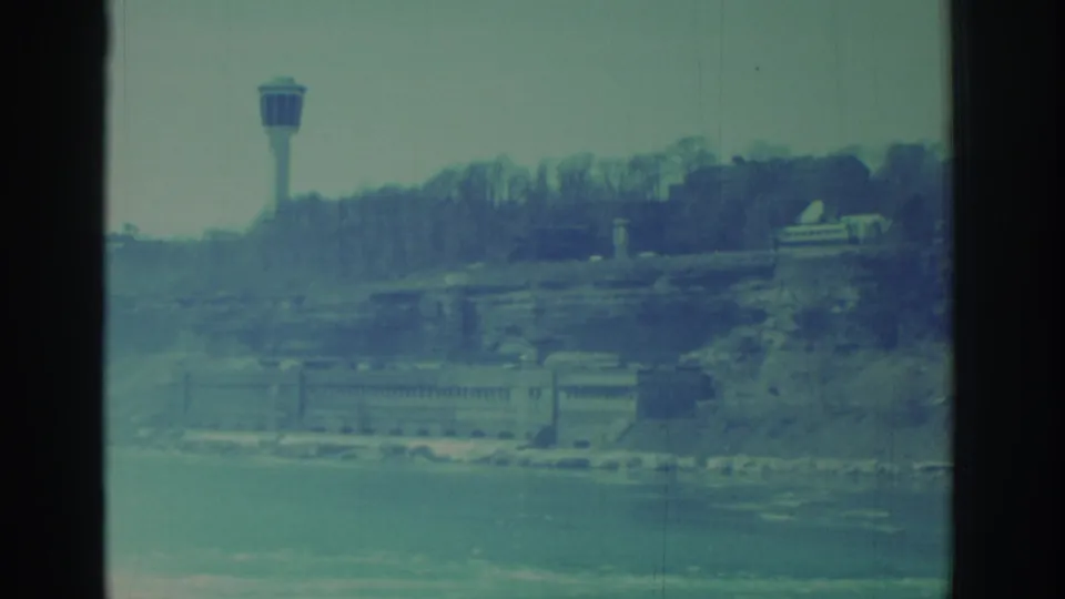 1971 8mm frame showing the Niagara Falls shoreline with a distant tower, bare trees, buildings, and water below.