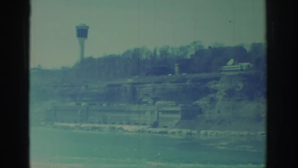Faded 1971 Niagara Falls home-movie frame with an observation tower, wooded cliff, riverside building, and water in the foreground.