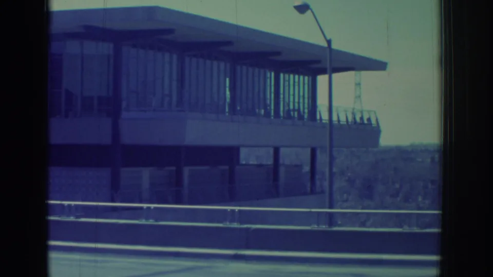 Final 1971 Niagara Falls-area frame showing the building's side elevation, roadside guardrails, a lamp post, and distant trees.