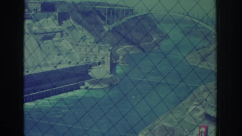 1971 visitor-shot 8mm view over Niagara Falls hydroelectric works, with river, bridge, shoreline, and safety fence visible.