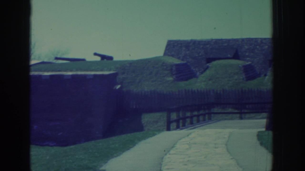 A historic fort approach in 1971 footage, with grassy ramparts, cannons, a path, and a wooden bridge seen through vertical framing.
