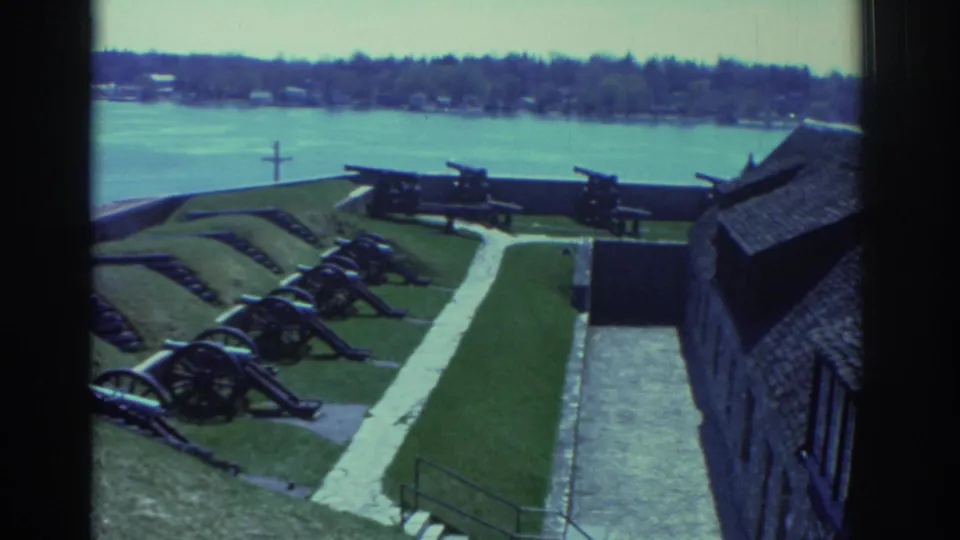 Visitor-shot 1971 view through a window frame across a historic fort's cannon display toward the river and distant shore.