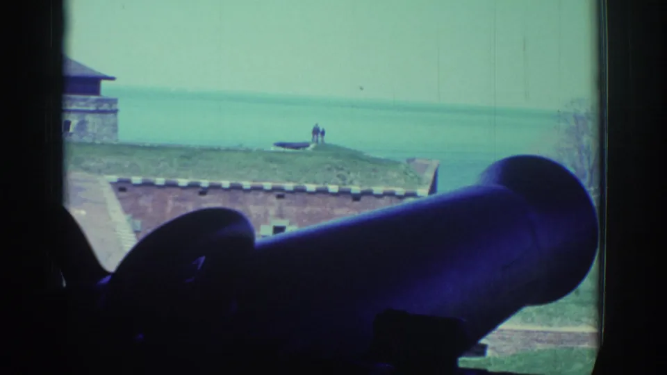 A large dark cannon or display piece frames a 1971 fort-window view toward the river, with tiny visitors visible near the water.