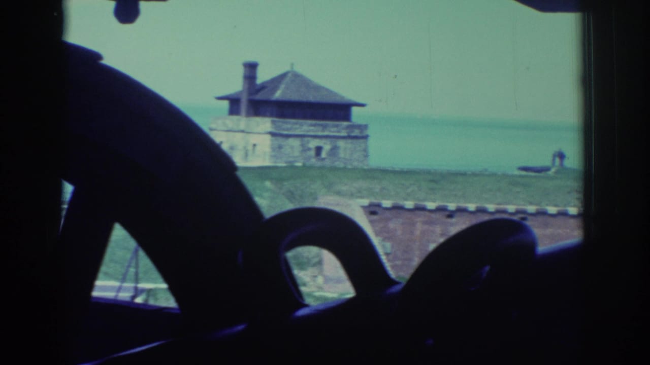 Visitor-shot 8mm frame of a historic fort near Niagara Falls, seen past dark cannon shapes toward a masonry building and water.