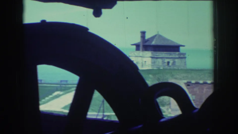 1971 home-movie frame looking from inside a fort opening past a cannon toward stonework, a path, and a waterfront building.