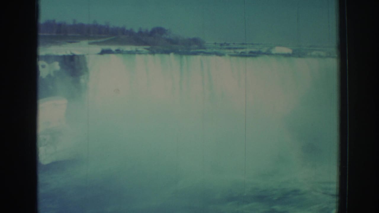 Wide waterfall view with heavy mist, distant shoreline, and vertical film scratches.