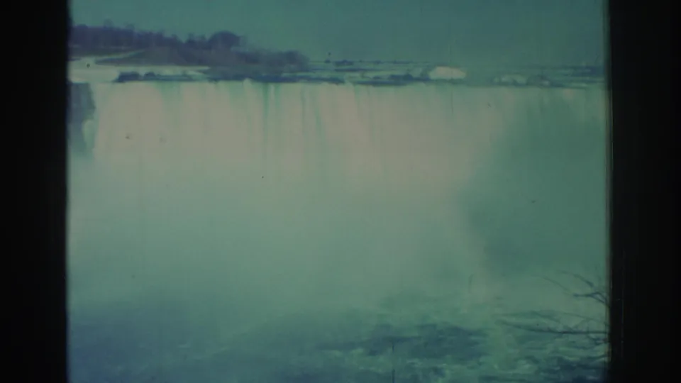 1971 8mm frame of Niagara Falls with a soft teal cast, dark contact-sheet divider, distant trees, and mist obscuring the base.