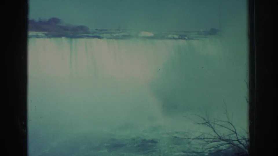 1971 Niagara Falls home-movie frame showing the falls through haze, with bare branches in the foreground and a distant shoreline above the water.