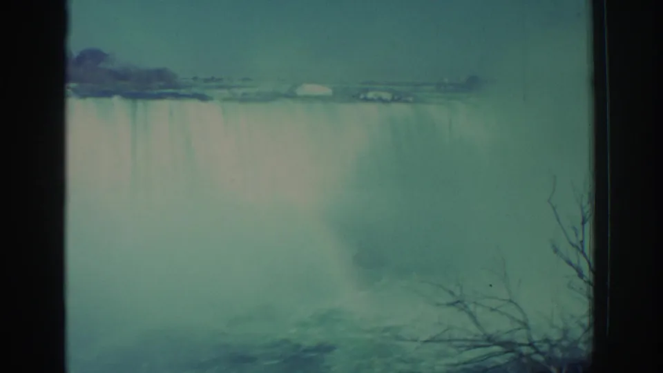 1971 8mm view of Niagara Falls with pale falling water, mist, teal sky, dark frame borders, and bare branches in the foreground.