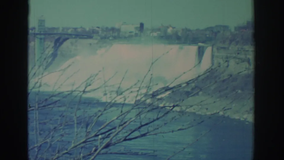 1971 8mm wide view of Niagara Falls with the city skyline, bridge structure, falling water, river below, and bare branches in front.