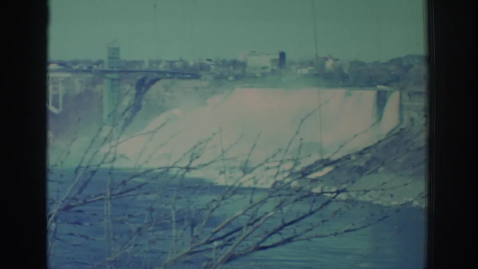 1971 Niagara Falls home-movie frame with a broad waterfall drop, mist, river below, distant skyline, bridge structure, and leafless branches.