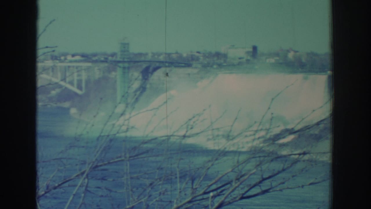Wide view of Niagara Falls mist, bridge structure, and bare branches in soft blue-green 8mm footage.