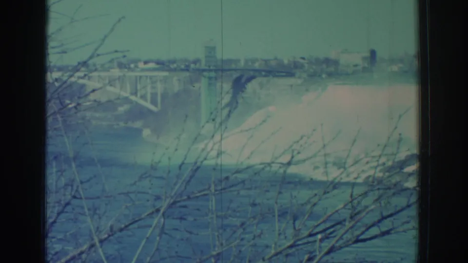 1971 Niagara Falls home-movie frame showing mist rising below a bridge or dam structure, with leafless branches in front.