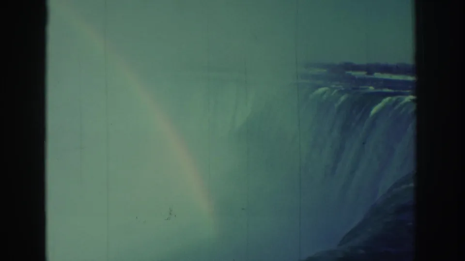 1971 home-movie frame showing rainbow color in the spray beside Niagara Falls, with grain, scratches, and a dark gate edge.