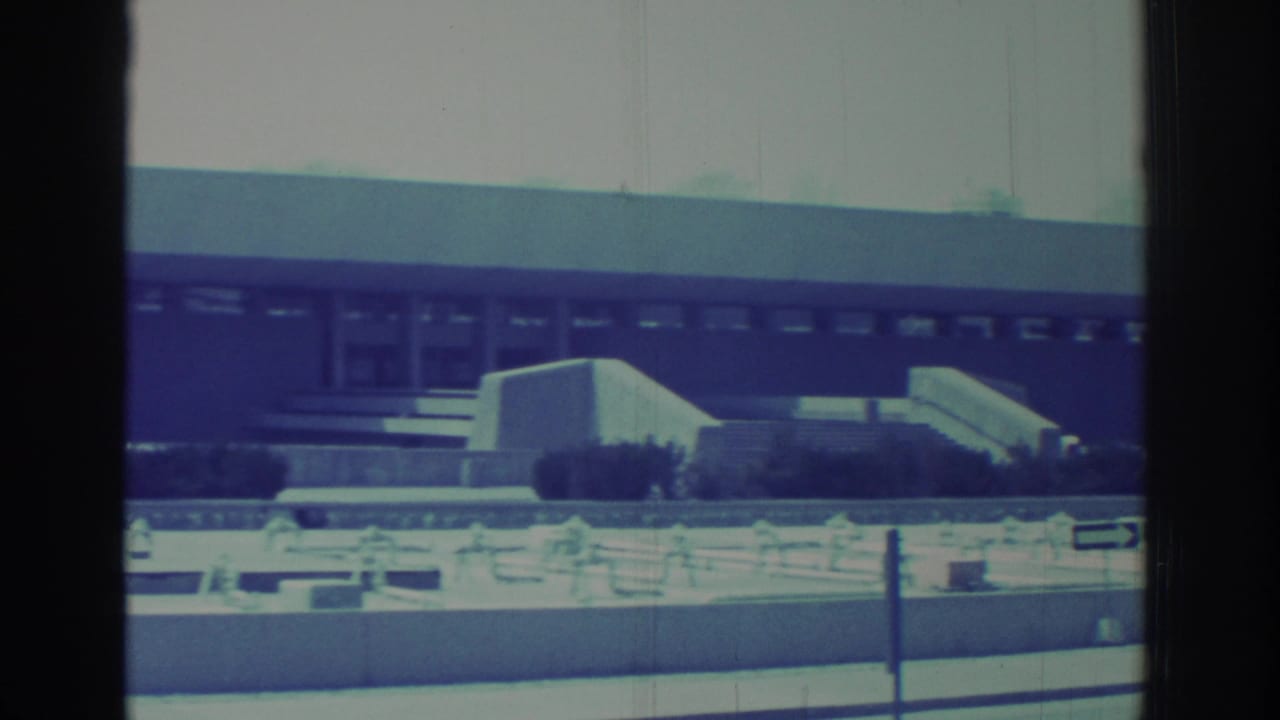 Long building facade with steps, windows, shrubs, and roadside foreground.