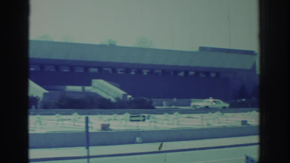 1971 8mm frame of the Hockey Hall of Fame building, roadside fence, entrance steps, shrubs, and a light-colored car in front.