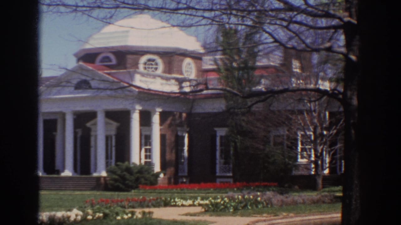 1971 8mm view of a brick domed mansion in Virginia, framed by bare branches, lawn, shrubs, and red spring flowerbeds.