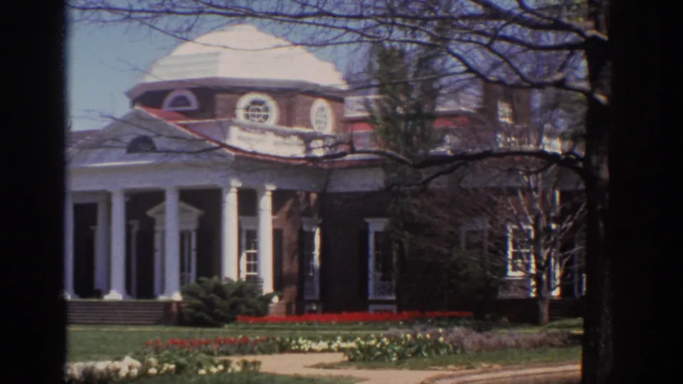 1971 8mm view of a brick domed mansion in Virginia, framed by bare branches, lawn, shrubs, and red spring flowerbeds.