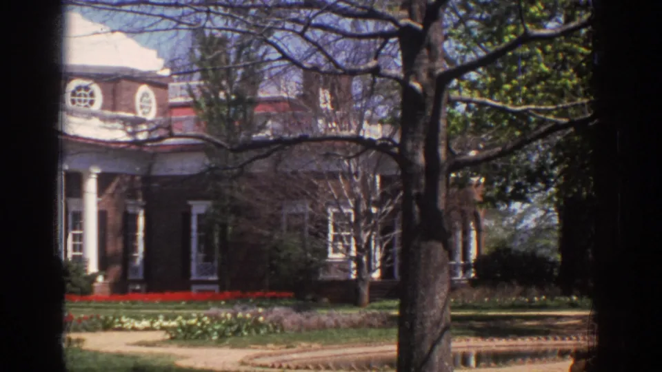 1971 8mm frame showing the mansion partly cropped, with brick walls, white columns, tree trunks, lawn, and red flowerbeds.