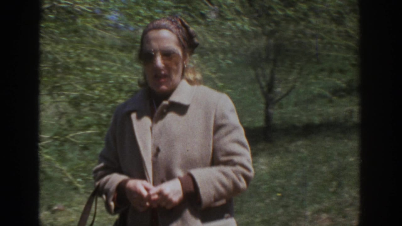 Woman in beige coat and sunglasses walking outdoors among green trees.