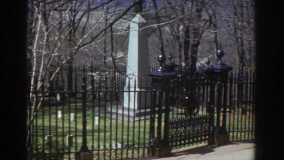 The handheld 8mm frame centers on a black iron gate and fence, with a pale stone obelisk visible behind bare branches and green grass.