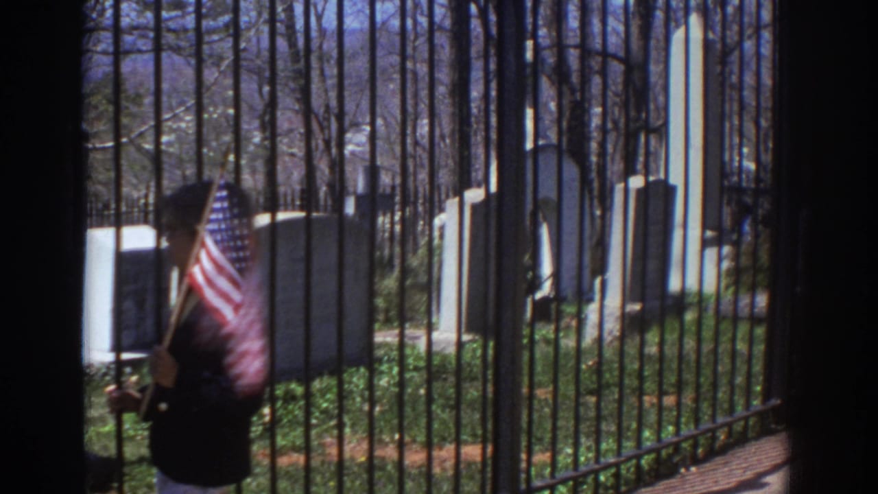1971 Richmond, Virginia 8mm frame showing a person with a small American flag near black iron cemetery bars, with upright headstones and bare trees beyond.