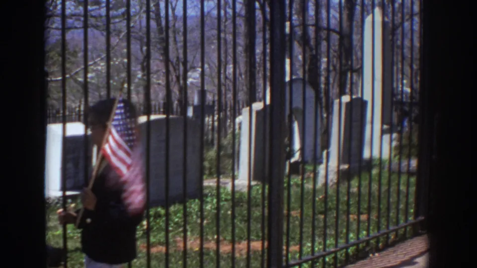 1971 Richmond, Virginia 8mm frame showing a person with a small American flag near black iron cemetery bars, with upright headstones and bare trees beyond.