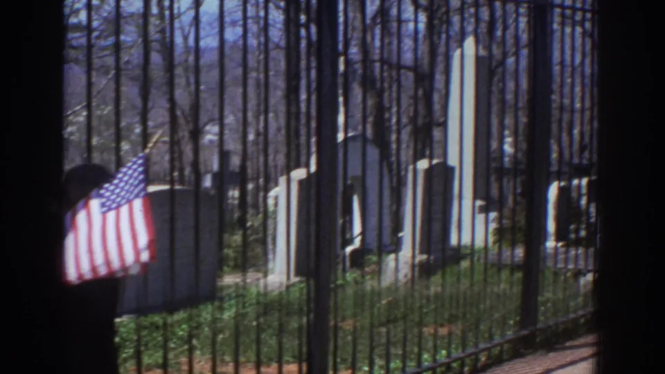 1971 Richmond, Virginia frame with a small American flag in front of a person at the cemetery fence, with monuments, an obelisk, and leafless trees in the background.