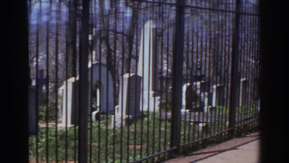 1971 Richmond, Virginia cemetery view through black iron fencing, with weathered headstones, a tall obelisk marker, bare trees, and ground cover inside the burial ground.