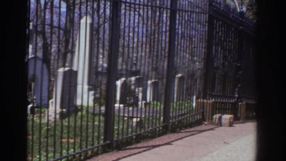 1971 Richmond, Virginia 8mm frame looking along an iron cemetery fence toward a walkway, headstones, a tall obelisk monument, bare trees, and sunlit ground.