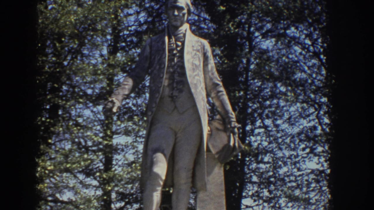 Full standing statue seen through trees in a sunlit park.