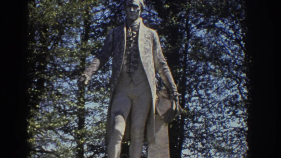 The James Monroe statue stands in a wooded Richmond park, with branches and strong sunlight surrounding the figure.