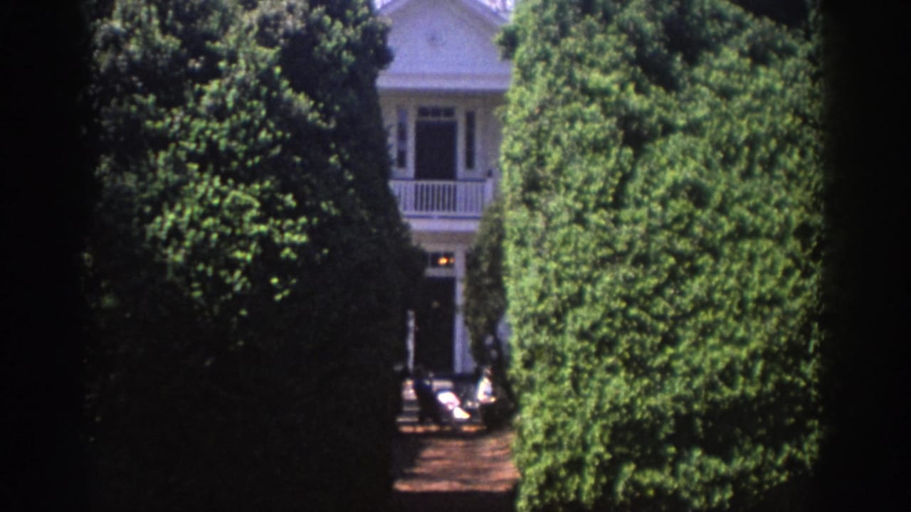 1971 Richmond home-movie frame showing a white house glimpsed between tall clipped hedges and trees.