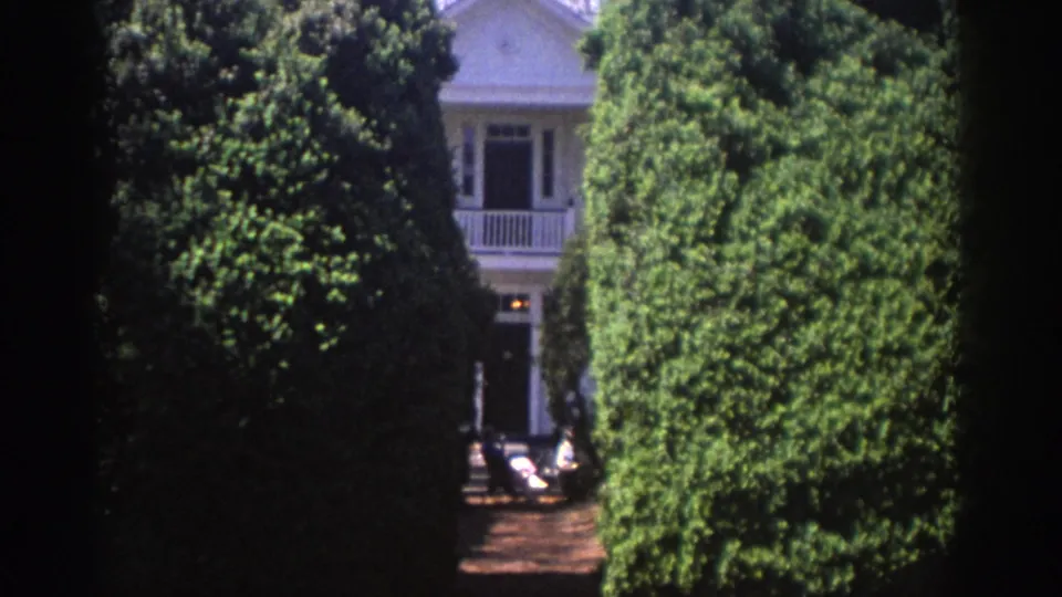 1971 Richmond home-movie frame showing a white house glimpsed between tall clipped hedges and trees.