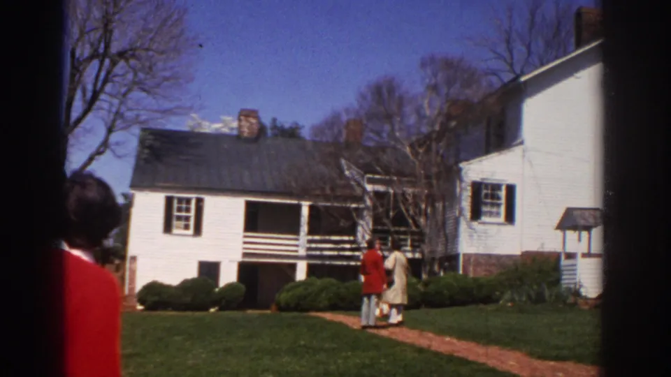 1971 Richmond frame with two adults on a grassy lawn near a white clapboard house and porch.