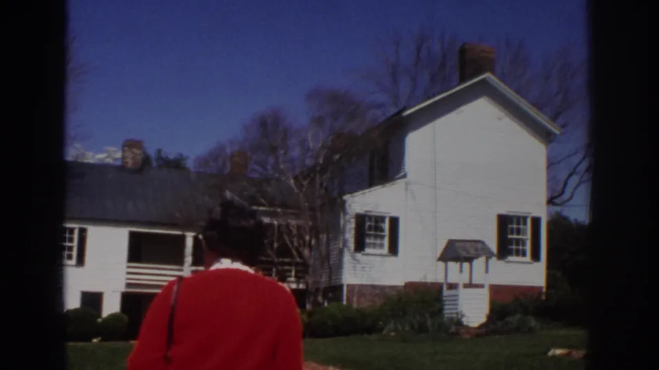 1971 Richmond home-movie frame showing a person in a red sweater from behind near a white house and landscaped yard.