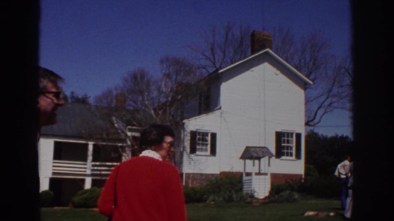 1971 Richmond home-movie frame showing adults walking near a white clapboard historic house with black shutters and bare trees.