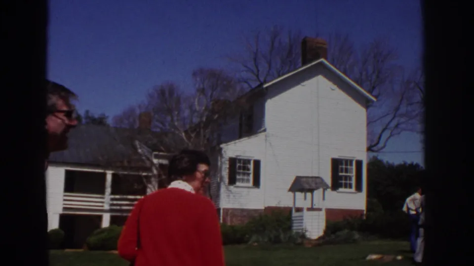 1971 Richmond home-movie frame showing adults walking near a white clapboard historic house with black shutters and bare trees.