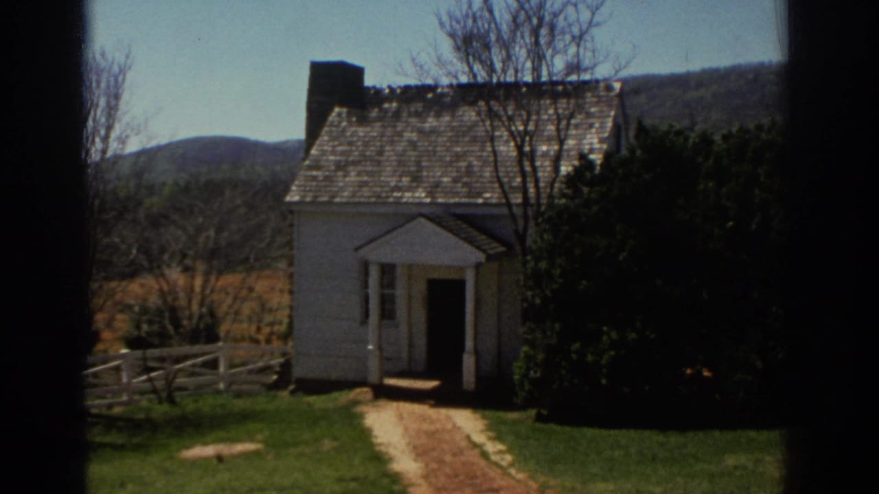 Small white house with a shingled roof, open doorway, fence, lawn, and hills behind it.