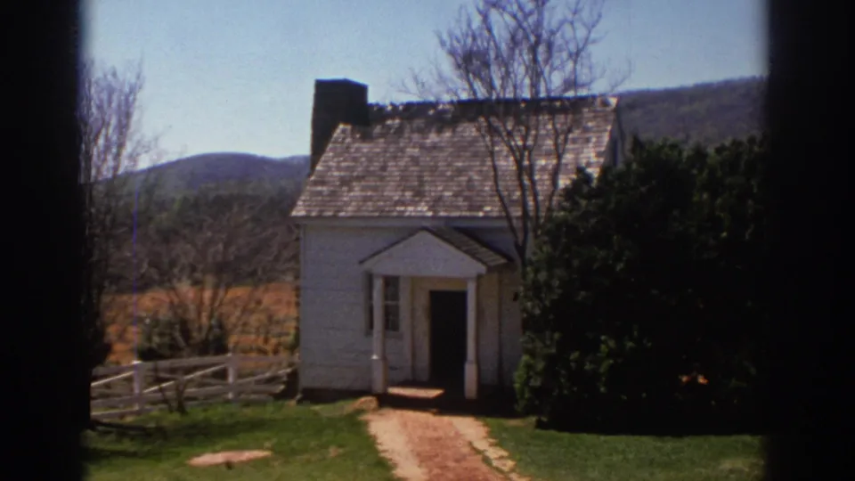 Handheld 1971 Richmond frame of a white historic house with a front path, split-rail fence, shrub, bare trees, and low hills.
