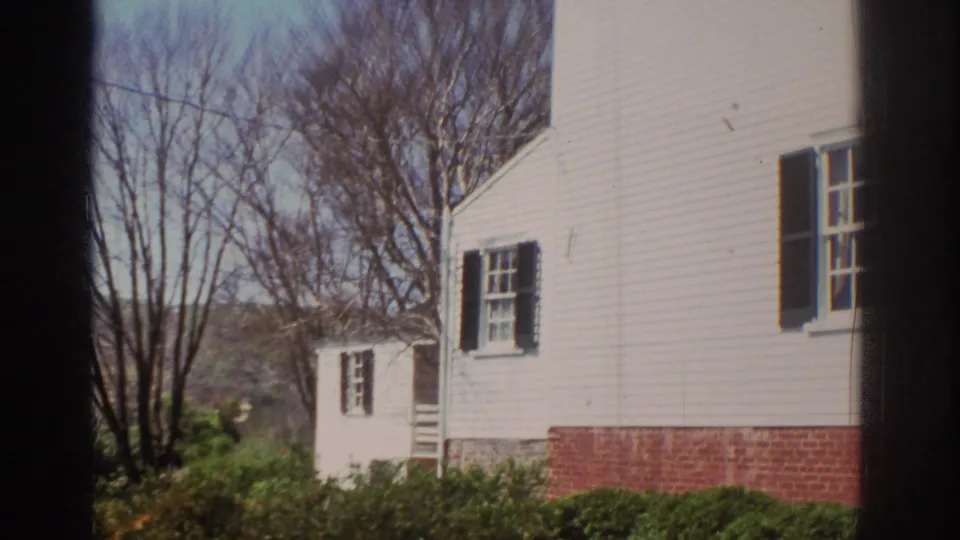 1971 Richmond frame showing the side wall of a white clapboard house with black shutters, brick foundation, shrubs, bare trees, and another small building.