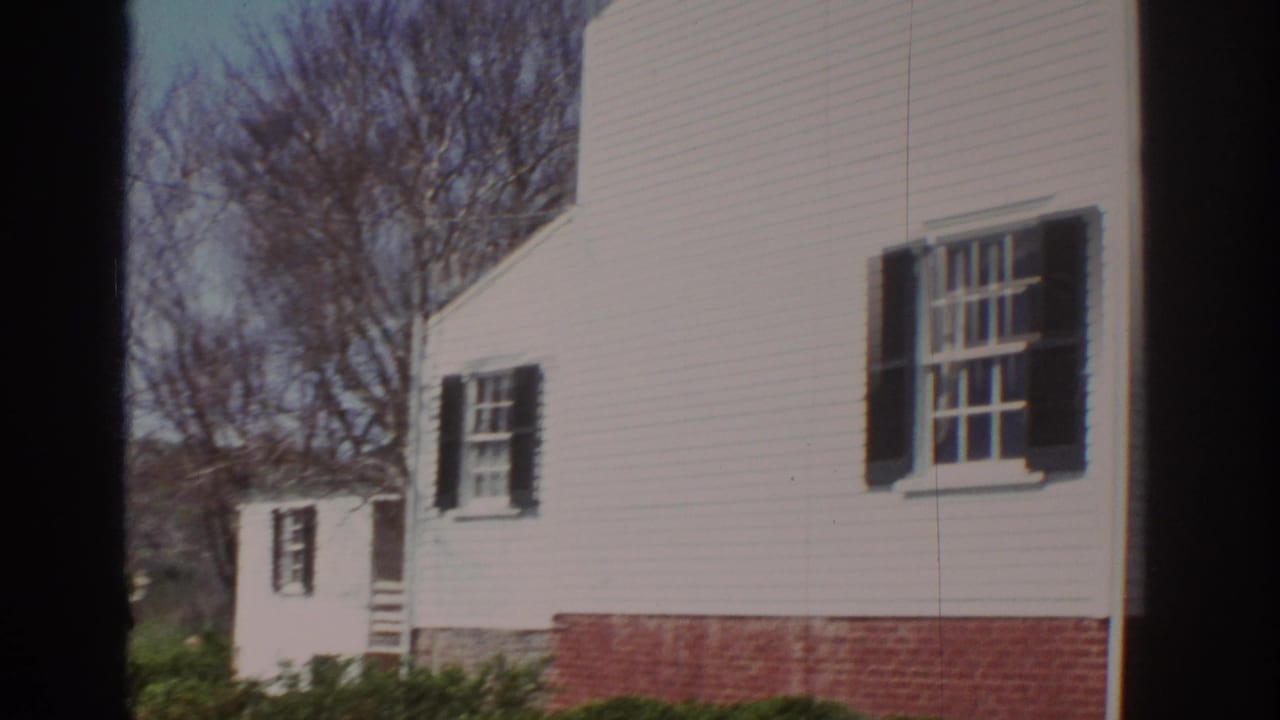 1971 Richmond home-movie frame showing a white clapboard house with black shutters, brick base, shrubs, and leafless trees.