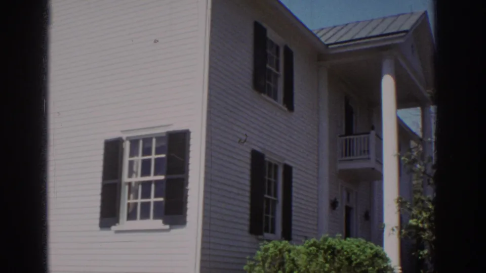 1971 Richmond frame showing a closer view of the house exterior, with shuttered windows, porch column, shrubs, and metal roof edge.