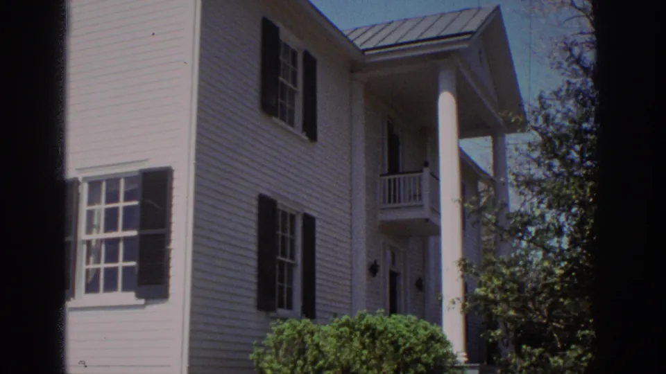 1971 Richmond home-movie frame of a white clapboard house facade with porch column, black shutters, metal roof, shrubs, and trees.