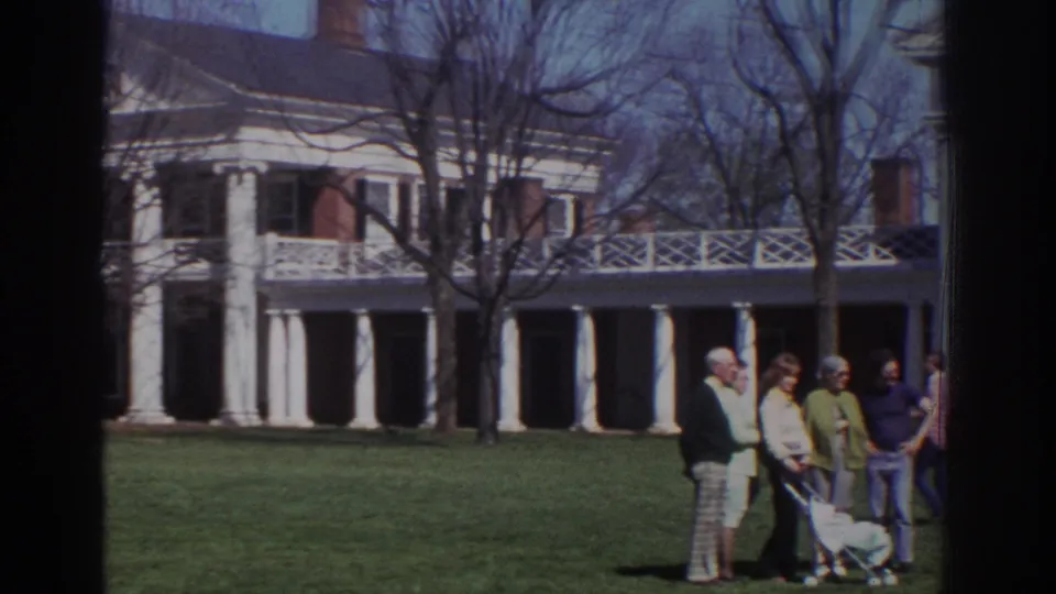 1971 Richmond home-movie frame with adults conversing beside a stroller on the lawn before a white-columned mansion.