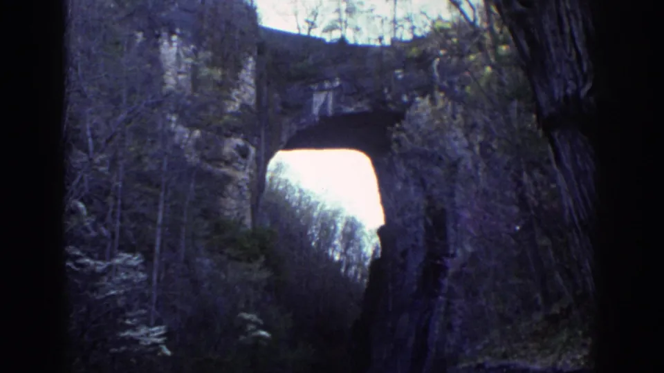 1971 home-movie frame showing the Natural Bridge opening centered between dark rock edges, trees, and forested limestone walls.