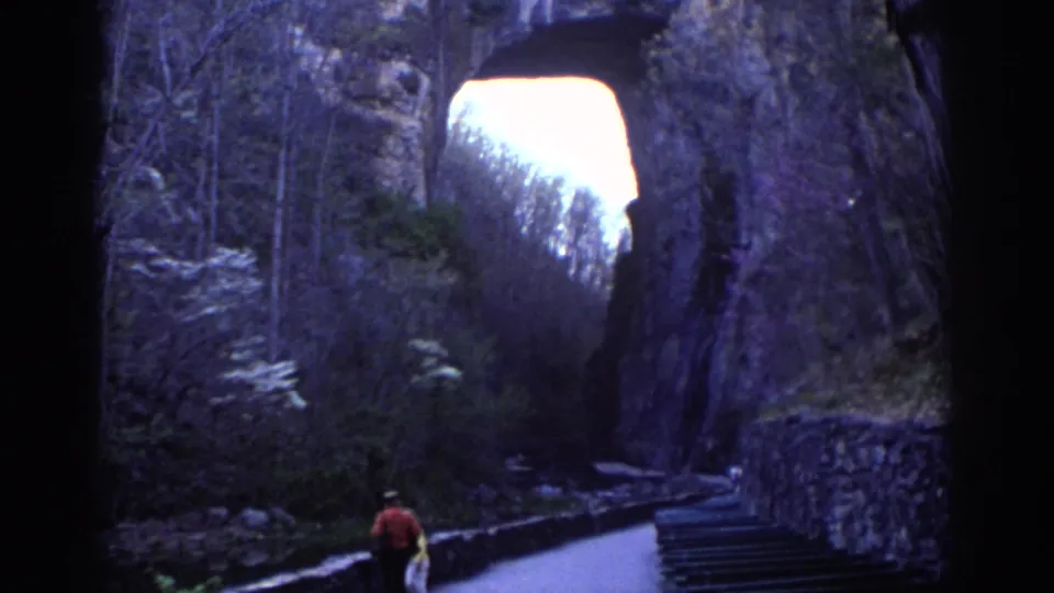 In the Richmond park scene, a distant pedestrian in red walks along a paved path toward the shadowed natural stone arch.