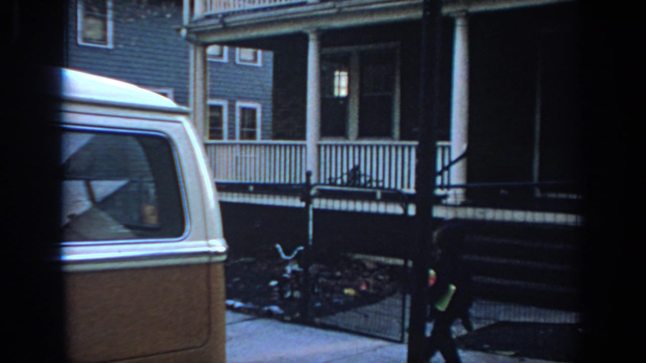 1972 Philadelphia home-movie frame with a two-tone van, porch railings, and a person nearing a front gate.